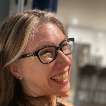 Woman wearing glasses smiling, side profile, indoors with blurred background.